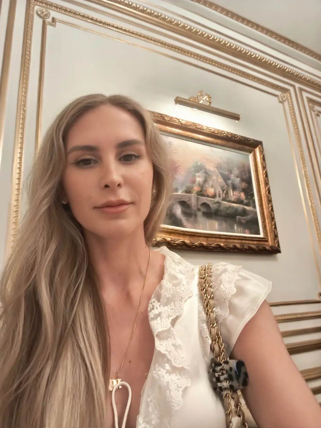 Isabelle Moreau Hamdan, with long fair hair, in a white lace blouse, standing in a room with gilded wall mouldings and a gilt-framed landscape painting behind her